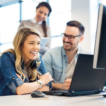 Junge Frau sitzt lachend vor Laptop im Büro mit zwei Kollegen